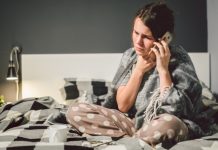 Vrouw zit op bed. Ze heeft een deken om zich heengeslagen en voelt aan haar keel. Ze is aan het bellen.